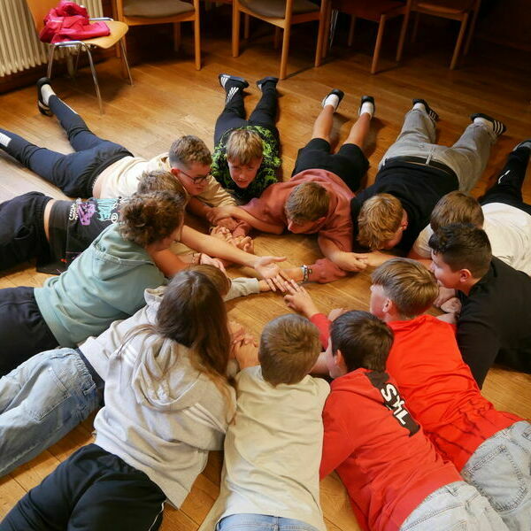 Kinder liegen auf dem Boden und spielen ein Kooperationsspiel, bei dem sie sich gegenseitig an den Armen festhalten.